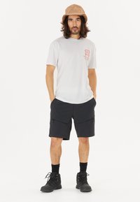 Vit kortärmad t-shirt med röd grafik, svarta shorts med dragkedjefickor och en ljusbrun bucket hat. Svarta stövlar fulländar outfiten.