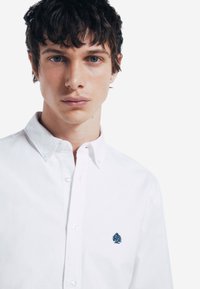 Camicia bianca con bottoni realizzata in cotone, caratterizzata da un colletto classico e un piccolo ricamo di un pino blu sul lato sinistro del petto.