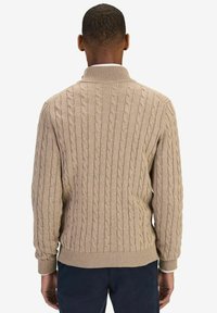 Beige kabelstickad tröja med hög krage, med ribbade ärmslut och nederkant, visad bakifrån. Slät textur med upphöjda mönster.