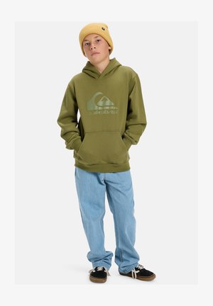 Ελαιόλαδο πράσινη hoodie με μπροστινή τσέπη καγκουρό και λογότυπο, συνδυασμένη με ανοιχτό μπλε τζιν και μαύρα αθλητικά παπούτσια με λευκές κορδόνες.