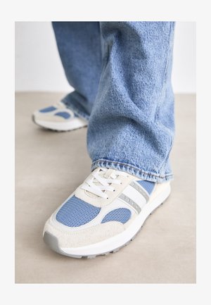 Sneakerit, joissa on vaaleanbeige ja sininen verkkovarsi, mokkanahkaosia, valkoiset nauhat ja harmaa kumipohja. Näkyvä brändäys sivussa.