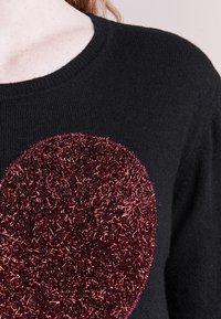 Pull noir en tricot avec un grand embellissement en forme de cœur composé de fibres métalliques bordeaux brillantes. Texture douce et col rond.