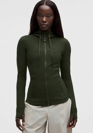 Mujer con una sudadera ajustada verde oscuro con cremallera y agujeros para el pulgar, y pantalones beige claros, de pie frente a un fondo gris liso.