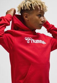 Roter Hoodie mit einer Fronttasche und weißem Logo. Das Material wirkt weich und hat eine glatte Textur. Verfügt über eine Kapuze mit Kordelzug und eng anliegende Ärmel.