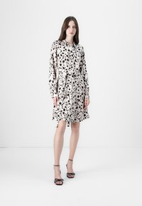 Beige kjole med svart prikket mønster, lange ermer, belted liv og en frynset skjørt. Kombinert med svarte høye sandaler med prikker.