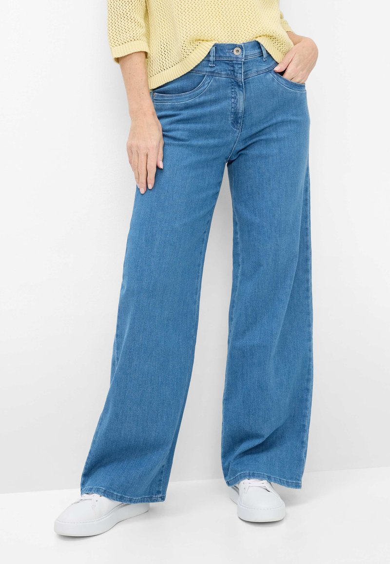 Person iført blå bukser med vide ben og forlommer, kombineret med hvide sneakers og en lys gul striktrøje.