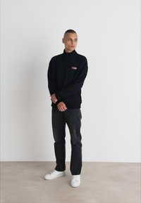 Melna zip-up kapuce ar diskrētu logo, kombinēta ar tumšiem džinsiem un baltām tenisēm. Gluda auduma un brīvi pieguļošs piegriezums. Minimālistisks dizains.