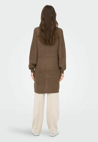 Maglione lungo marrone a maglia con un motivo testurizzato, maniche oversize e polsini a coste. Abbinato a pantaloni crema a gamba larga e scarpe bianche.