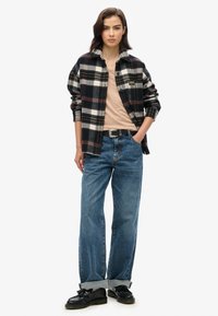 Camisa de franela a cuadros en tonos negro, blanco y burdeos sobre una parte superior de color beige claro, combinada con unos vaqueros de pierna ancha de color azul claro y zapatos negros.
