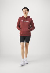 Bordó kapucnis pulóver elülső zsebbel, fehér logós szöveggel. Fekete, sima felszínű rövidnadrág. Fehér sportcipő és bokaszalag. 