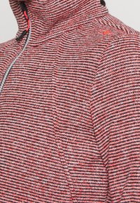 Veste en polaire à motif rouge et noir avec un col montant, une fermeture éclair frontale et un tissu texturé, arborant un design sportif.