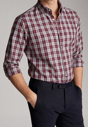 Mann trägt burgunderrote und graue karierte Hemd mit hochgekrempelten Ärmeln, in schwarze Hose gesteckt, Hände in den Taschen.