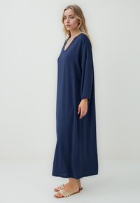 Abito lungo navy con scollatura a V profonda, maniche lunghe e vestibilità ampia. Realizzato in tessuto morbido. Indossato con sandali a strappo di colore chiaro.