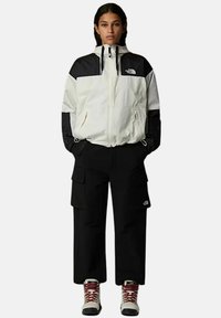 Biała i czarna kurtka z zapięciem na zamek, dwiema kieszeniami i regulowanym sznurkiem. Czarne spodnie cargo z kieszeniami bocznymi. Białe buty z czerwonymi sznurowadłami.