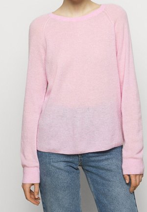 Personne portant un pull à manches longues rose clair et un jean en denim bleu, debout les bras détendus le long du corps.