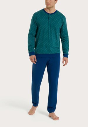 Uomo che indossa una maglia da pigiama a maniche lunghe color verde acqua con piccoli puntini e polsini e colletto blu scuro, abbinata a pantaloni da pigiama blu scuro, a piedi nudi.