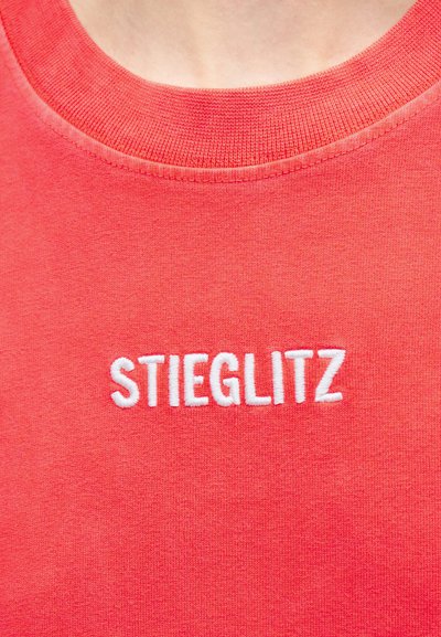 Κόκκινο πουκάμισο με λευκή κεντημένη επιγραφή "STIEGLITZ" κεντραρισμένη κάτω από το λαιμό στην περιοχή του στήθους.