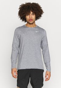 Camiseta deportiva de manga larga en tejido gris con un cuello redondo. Presenta un pequeño logo blanco de Nike en el pecho y un ajuste relajado.