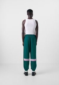 Calças leves verdes com riscas horizontais rosas na parte inferior das pernas, cós elástico e um corte casual. Usadas com uma t-shirt sem mangas branca.