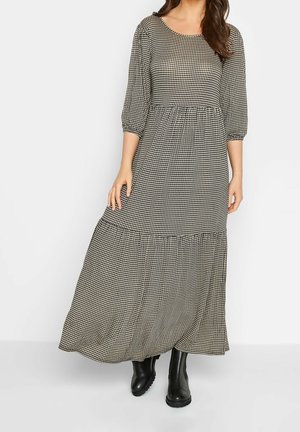 Kobieta w długiej sukience z drobnym czarno-białym wzorem pepitki, z rękawami do połowy przedramienia, nosząca czarne botki za kostkę.