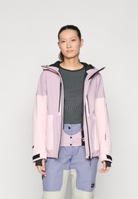 Lys rosa vanntett jakke med svarte detaljer, glidelåslommer og hette, båret over en mørkegrå topp og flerfargede bukser. Glatt tekstur.