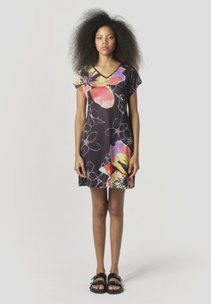 Czarna sukienka z żywym kwiatowym wzorem w kolorach czerwonym, żółtym i niebieskim, z dekoltem w kształcie litery V i krótkimi rękawkami typu cap. Noszona z czarnymi sandałami.