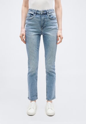 Τζιν στενή εφαρμογή σε ανοιχτό μπλε χρώμα, φορεμένο με λευκά αθλητικά παπούτσια, εμφανίζεται από τη μέση ως τα πόδια μπροστά σε απλό λευκό φόντο.