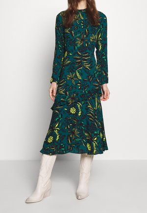 Vrouw draagt een lange mouwen teal bloemenjurk met ruchtdetail, gecombineerd met kniehoog witte laarzen tegen een effen achtergrond.