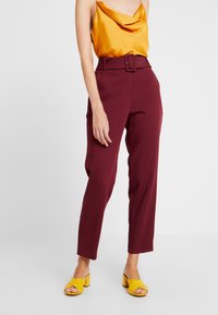 Maroon high-waisted trousers with a wide belt, paired with a draped yellow satin top. Worn with yellow block-heeled sandals.