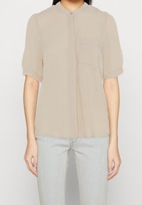Blusa beige a maniche corte con colletto rotondo, taschino singolo, tessuto morbido e chiusura con bottoni. Materiale leggero con vestibilità ampia.
