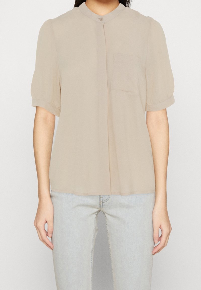 Blusa beige a maniche corte con colletto rotondo, taschino singolo, tessuto morbido e chiusura con bottoni. Materiale leggero con vestibilità ampia.