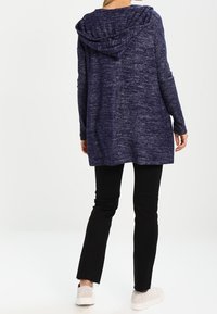 Felpa con cappuccio lunga di colore blu navy, con un motivo texturizzato e mélange, dotata di cappuccio con coulisse, vestibilità comoda e tasche laterali, abbinata a pantaloni neri.