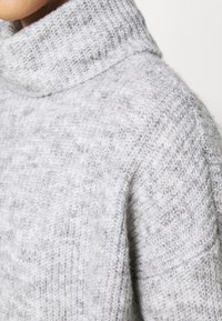 Pull gris en tricot avec un col montant côtelé. Le matériau semble doux et duveteux, avec un motif texturé et une coupe légèrement ample.