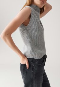 Eine Frau trägt einen grauen, ärmellosen, gerippten Rollkragenpullover und schwarze Jeans, mit einer Hand in ihrer Gesäßtasche.