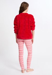 Červený fleecový pulovr s texturovaným vzorem srdce. V kombinaci s bílými legínami s červeným a růžovým geometrickým vzorem. Modelka je naboso.