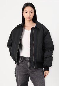 Veste bomber noire avec fermeture éclair à l'avant, grand col et poignets côtelés. Portée par-dessus une chemise blanche et associée à un jean foncé. Texture lisse.