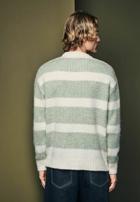 Striped sweater featuring green and white colors, knitted texture, crew neckline, and ribbed cuffs, paired with dark denim jeans.