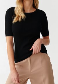 Schwarzes geripptes Strickoberteil mit kurzen Ärmeln, Rundhalsausschnitt und figurbetonter Silhouette, kombiniert mit hellbeigen Hosen.