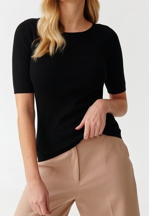 Schwarzes geripptes Strickoberteil mit kurzen Ärmeln, Rundhalsausschnitt und figurbetonter Silhouette, kombiniert mit hellbeigen Hosen.