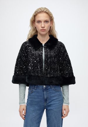 Le mannequin porte une veste noire à sequins, avec un col et des poignets moelleux, sur un haut gris et un jean bleu, devant un fond uni.