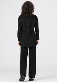 Svart kappa med midjebälte, långa ärmar och knappade manschetter, i kombination med matchande svarta vida byxor och ankelskor.