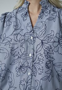 Knapbluse i let stof med blå og hvide striber, der har blomsteromrids, ruffles ved kraven og knapper foran.