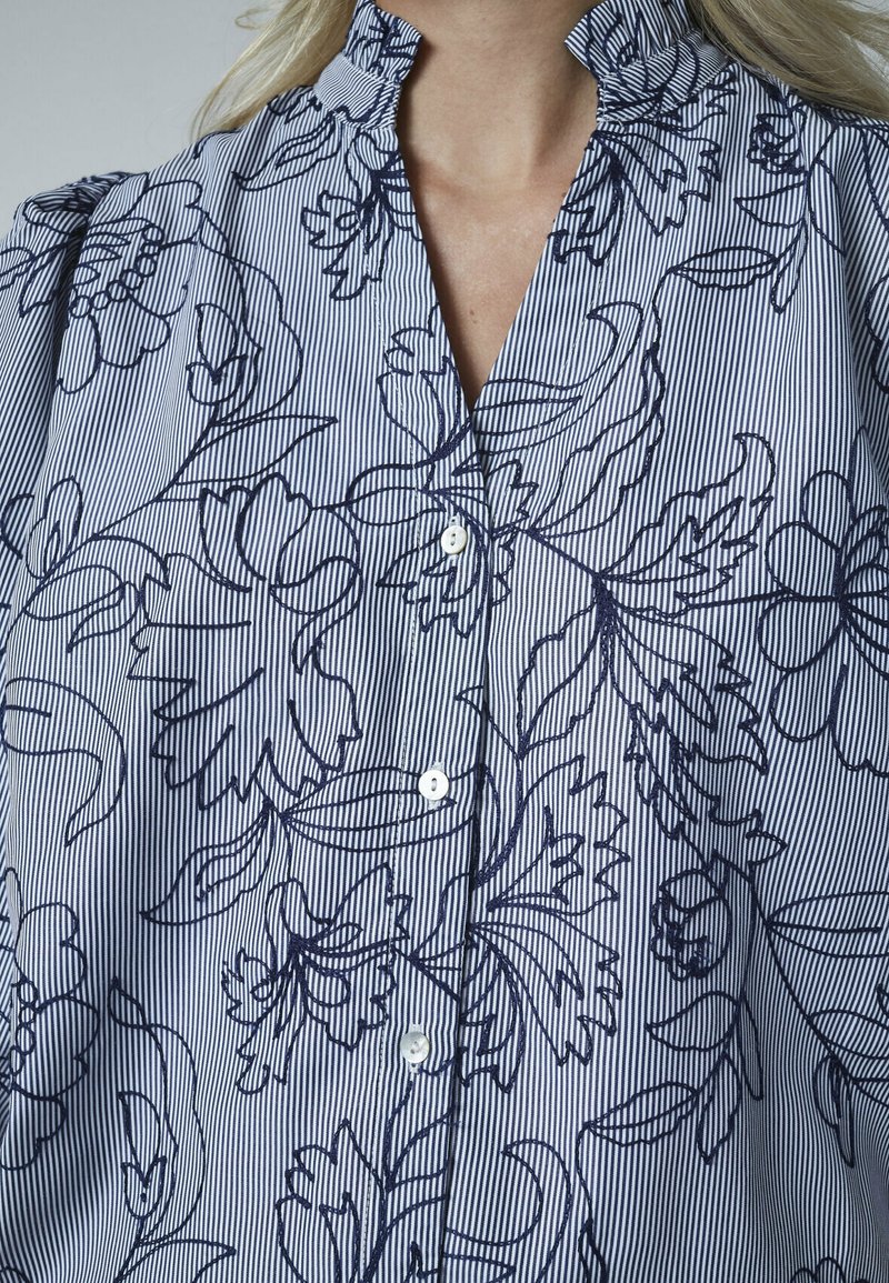 Knapbluse i let stof med blå og hvide striber, der har blomsteromrids, ruffles ved kraven og knapper foran.