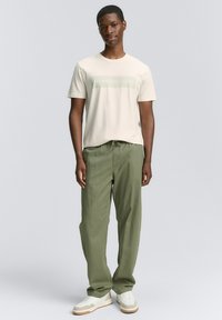 Camiseta de algodón beige con logo verde en la parte frontal, combinada con pantalones holgados de color verde oliva y zapatillas blancas con detalles marrones.