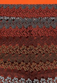 Flerlagers tyg med texturerad virkdesign i nyanser av röd, brun och svart, med zickzackmönster och en ribbad orange topp.