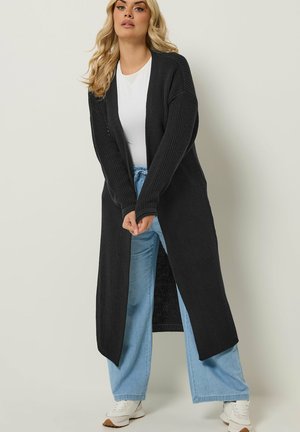 Kvinde iført en lang sort strikket cardigan over en hvid skjorte, lyseblå vidde jeans og hvide sneakers mod en ensfarvet baggrund.