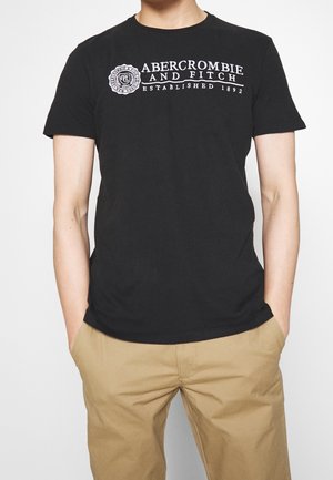 Osoba nosząca czarną koszulkę Abercrombie and Fitch i beżowe spodnie, z rękami w kieszeniach, na białym tle.