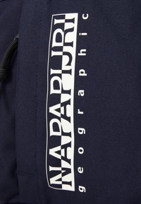 Marinblå tygryggsäck med en stor vit logotyp "NAPAPIJRI" och ordet "geographic". Har en texturerad yta och dragsnöre.