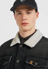 Ung man som bär en svart keps och en svart denimjacka med vit sherpa-krage, neutral uppsyn, vänd mot kameran på en enfärgad bakgrund.