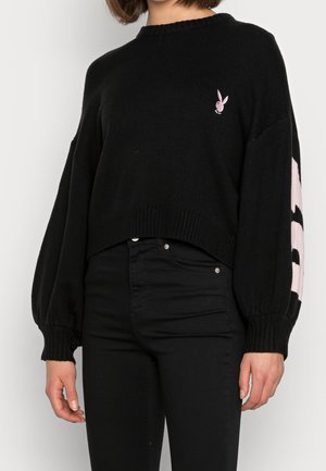 Kvinde iført en sort cropped sweater med puffede ærmer, pink logo på brystet og sorte højtaljede bukser mod en ensfarvet baggrund.